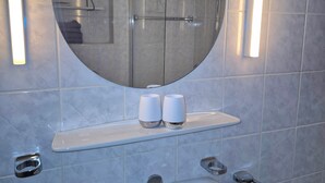 Une salle de bain avec un miroir rond, un lavabo, deux tasses posées sur une étagère et un porte-serviettes.