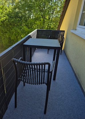 Ein Balkon mit Tisch und zwei Stühlen, Blick auf Grünflächen und eine Gebäudefassade.