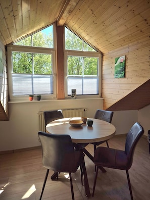 Ein Zimmer mit einem runden Tisch, vier Stühlen, zwei Fenstern mit Jalousien und einem Heizkörper.
