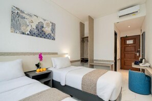 Une chambre d’hôtel avec deux lits, une table de chevet ornée d’un vase et de quelques oranges, une armoire en bois et une porte de salle de bain.