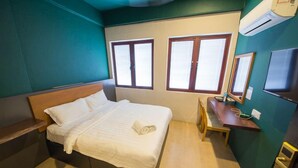 Un letto singolo con testiera in legno, un comodino e una sedia. Sono presenti due finestre con stipiti in legno e tende bianche.