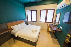 Ein Einzelbett mit Holzüberbau, ein Nachttisch und ein Stuhl. Zwei Fenster mit Holzrahmen und weißen Vorhängen sind zu sehen.
