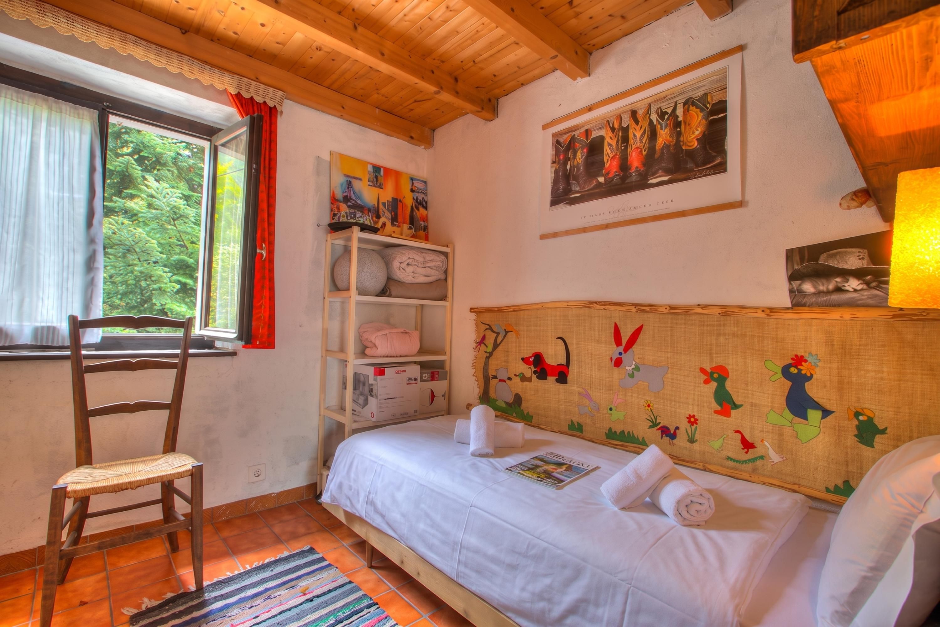 A bedroom with a wooden ceiling, a bed with white bedding, a chair, a window with a view of trees, and a shelf with items.