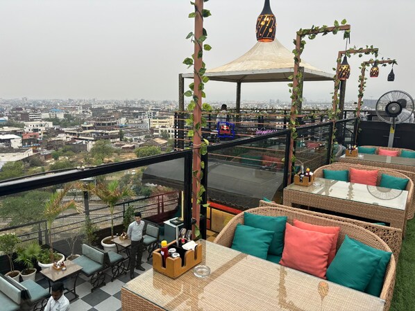 A rooftop bar with wicker furniture, a glass table, and a view of the cityscape. - Hotel Grand Sky (Jaipur)