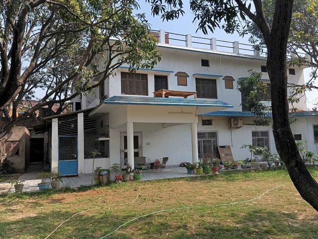 A two-story building with a covered porch, a small garden area with potted plants, and a tree in the foreground.