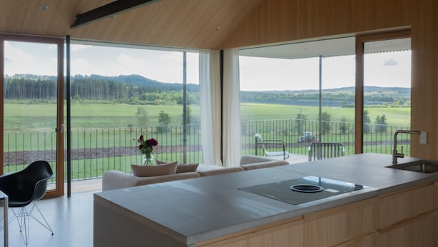 A modern kitchen with a large island, a dining area with a sofa and chairs, and a view of a green landscape through large windows. - Apartment 'Ruhige Dachgeschoss-Wohnung' with Panoramic Green View, Balcony and Parking (Bad Dürrheim)