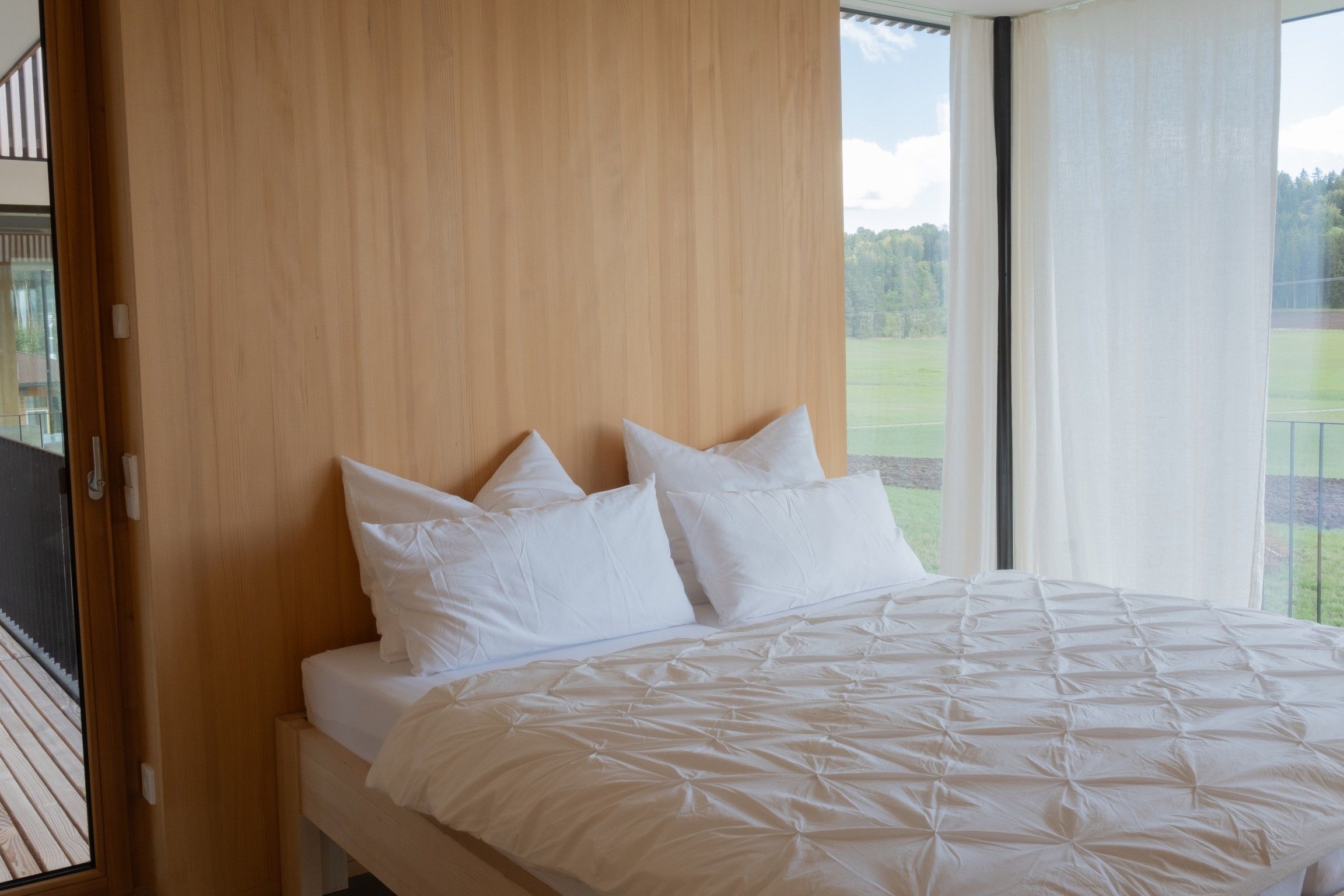 A bedroom with a wooden headboard, a large bed with white bedding, and a view of a green landscape through a window.