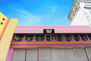 A hotel with a pink and yellow facade, a signboard with the hotel name, and closed shutters.