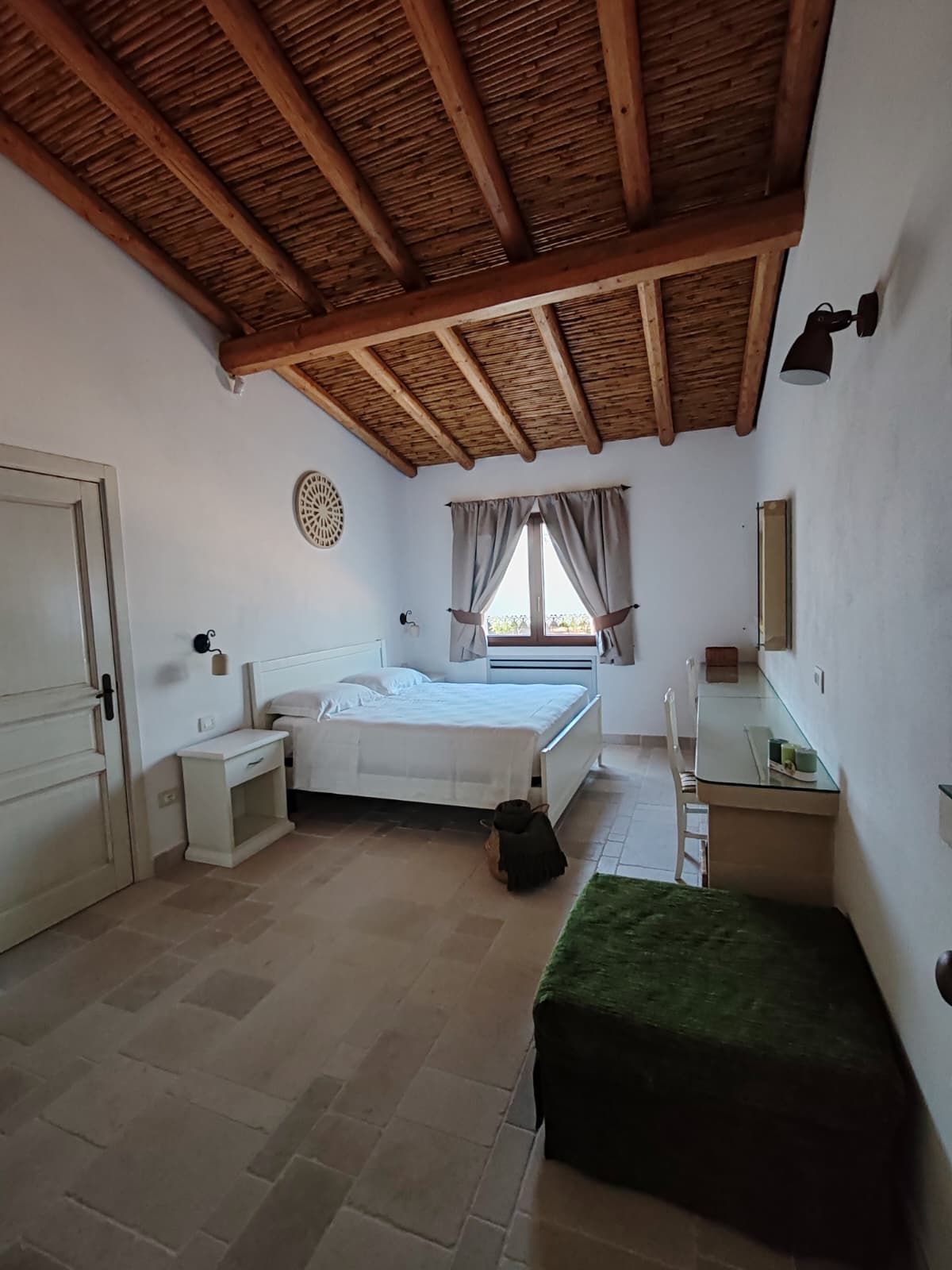 A bedroom with a bed, bedside tables, a small bench, a window with curtains, and a ceiling with wooden beams.