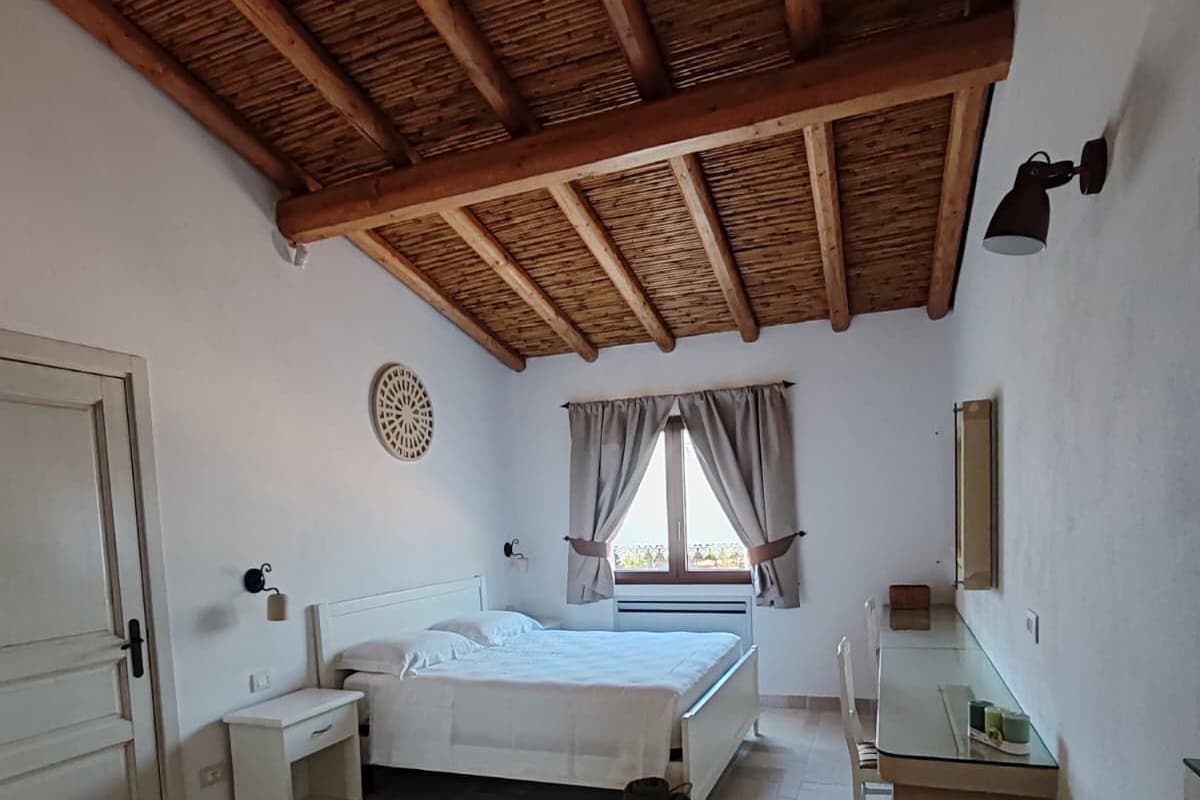 Ein Schlafzimmer mit einem Bett, Nachttischen, einer kleinen Bank, einem Fenster mit Vorhängen und einer Holzbalkendecke.