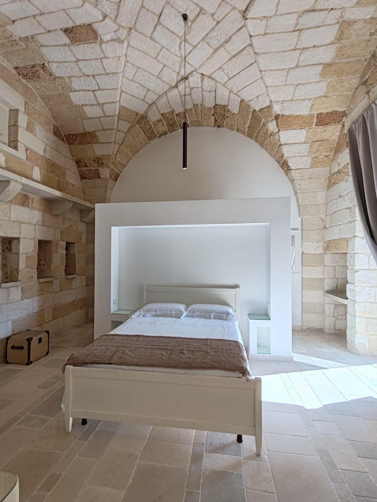 Un dormitorio con pared de piedra, cama blanca y lámpara colgante.