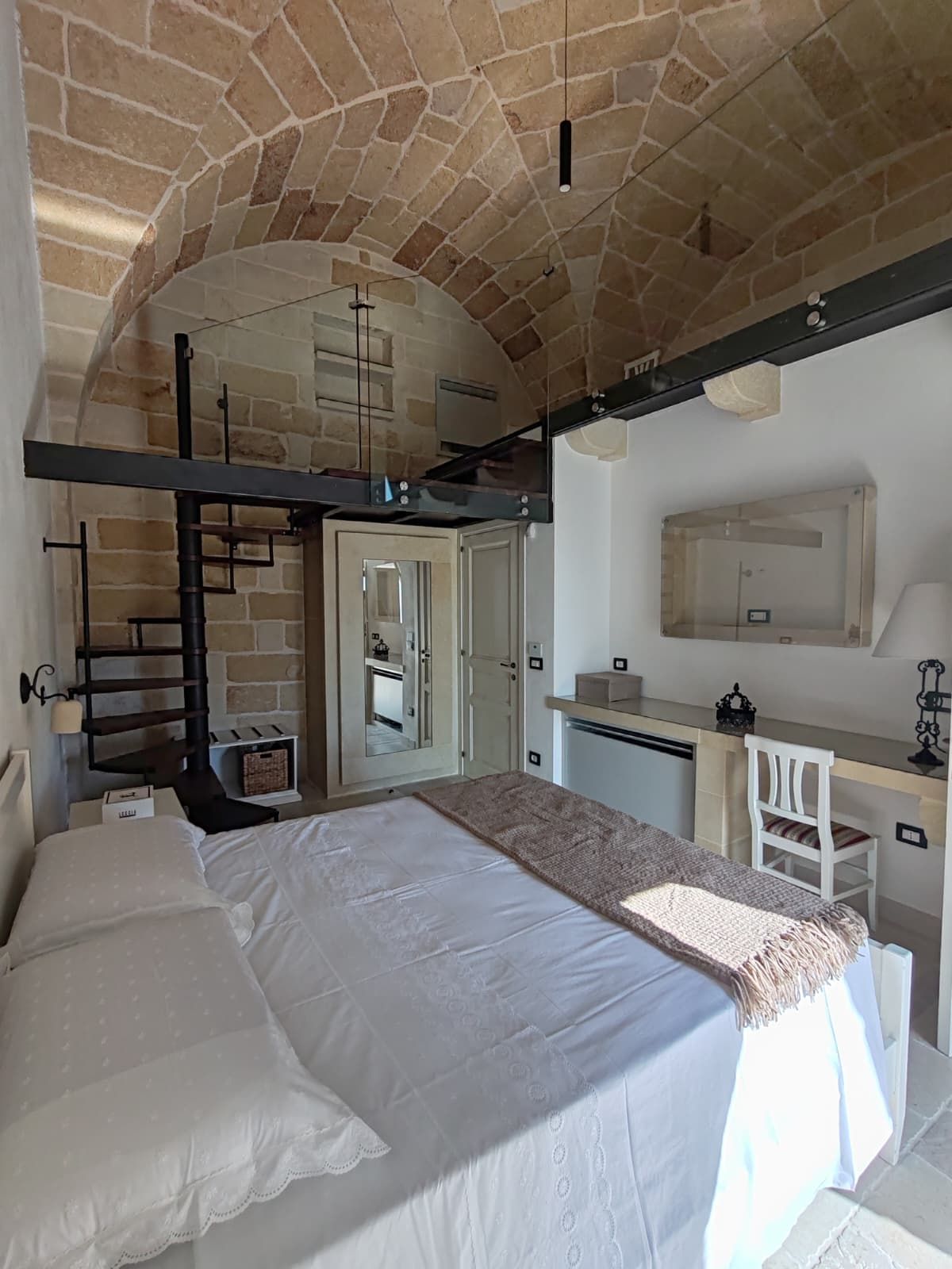 Un dormitorio con techo de piedra, cama con ropa de cama blanca, escritorio con silla y espejo.