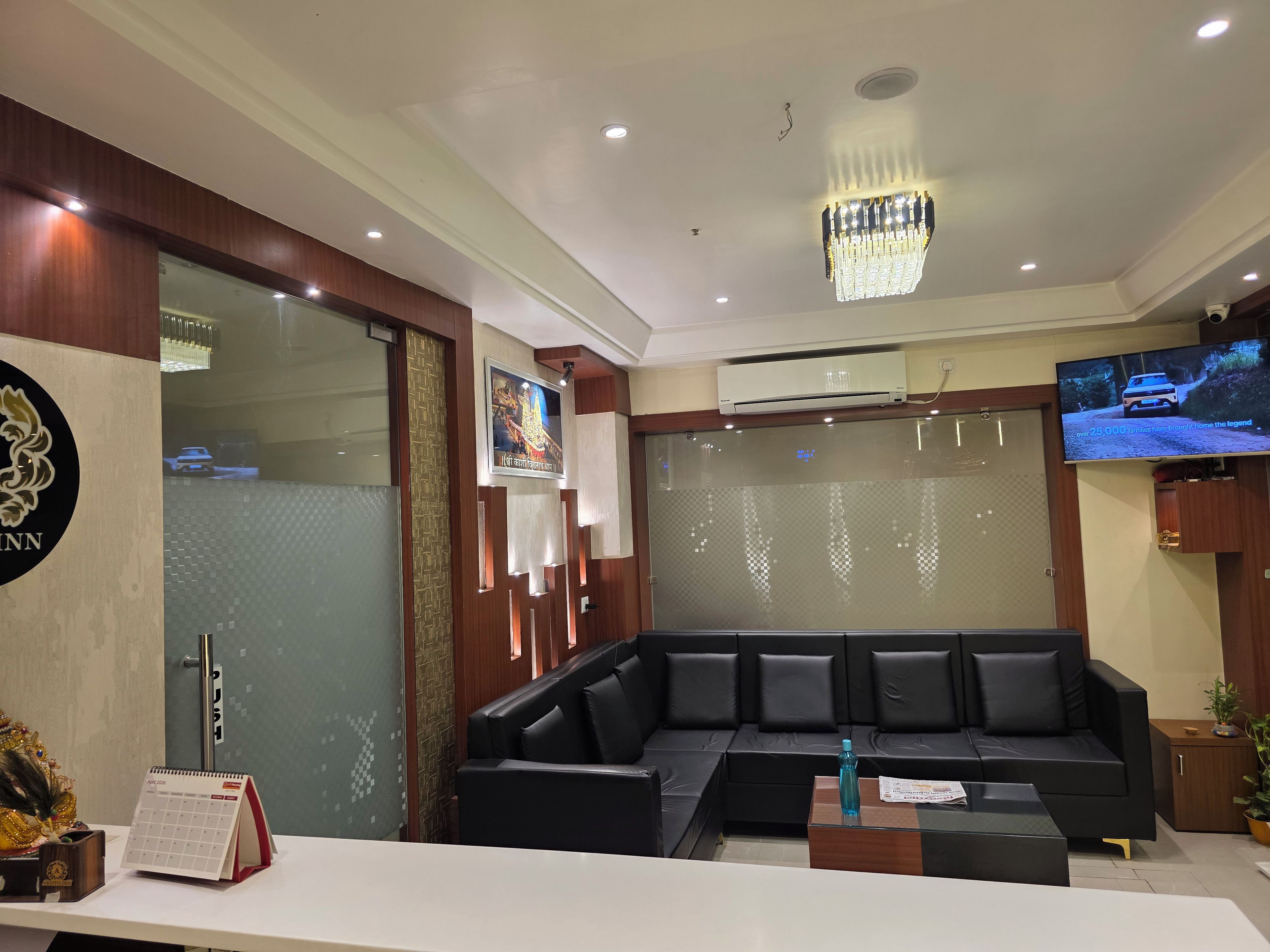 A modern reception area with a black leather sofa, a wooden reception desk, a mounted television, and a chandelier.