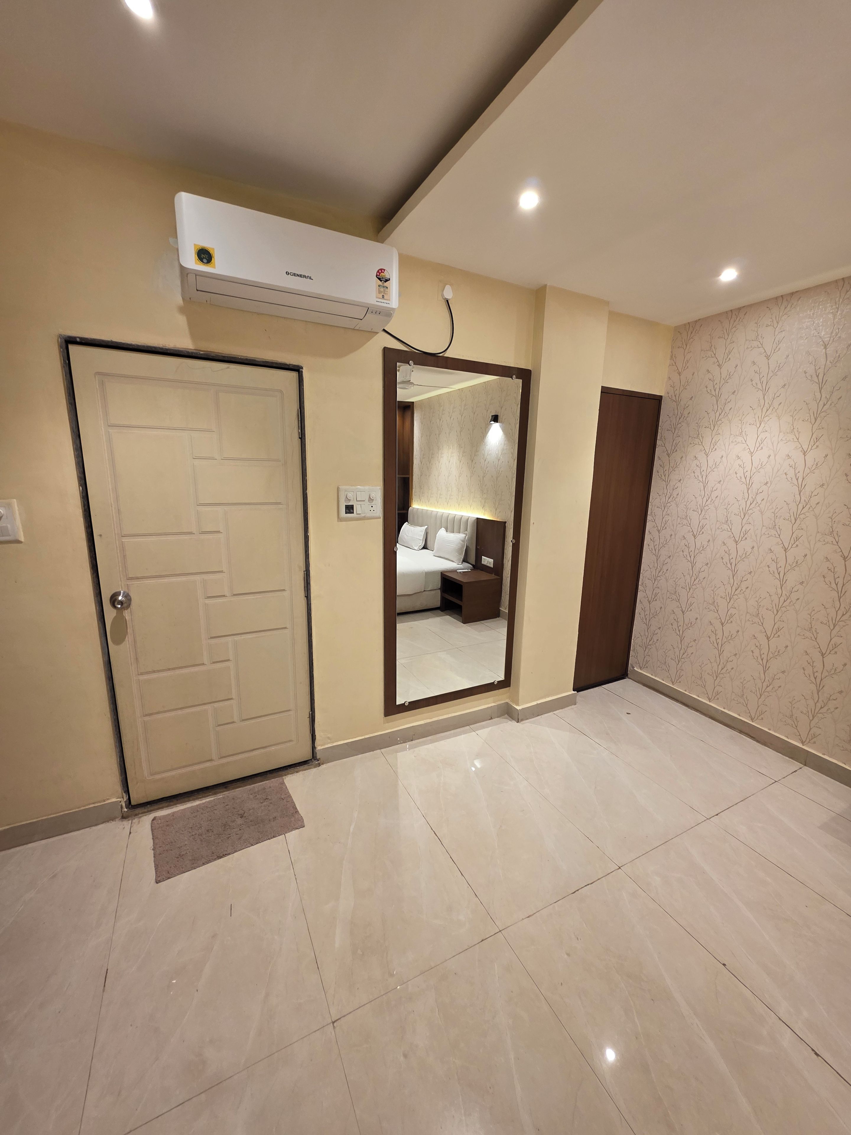 A hotel room with a bed, bedside table, and a large mirror. There is an air conditioning unit mounted on the wall.