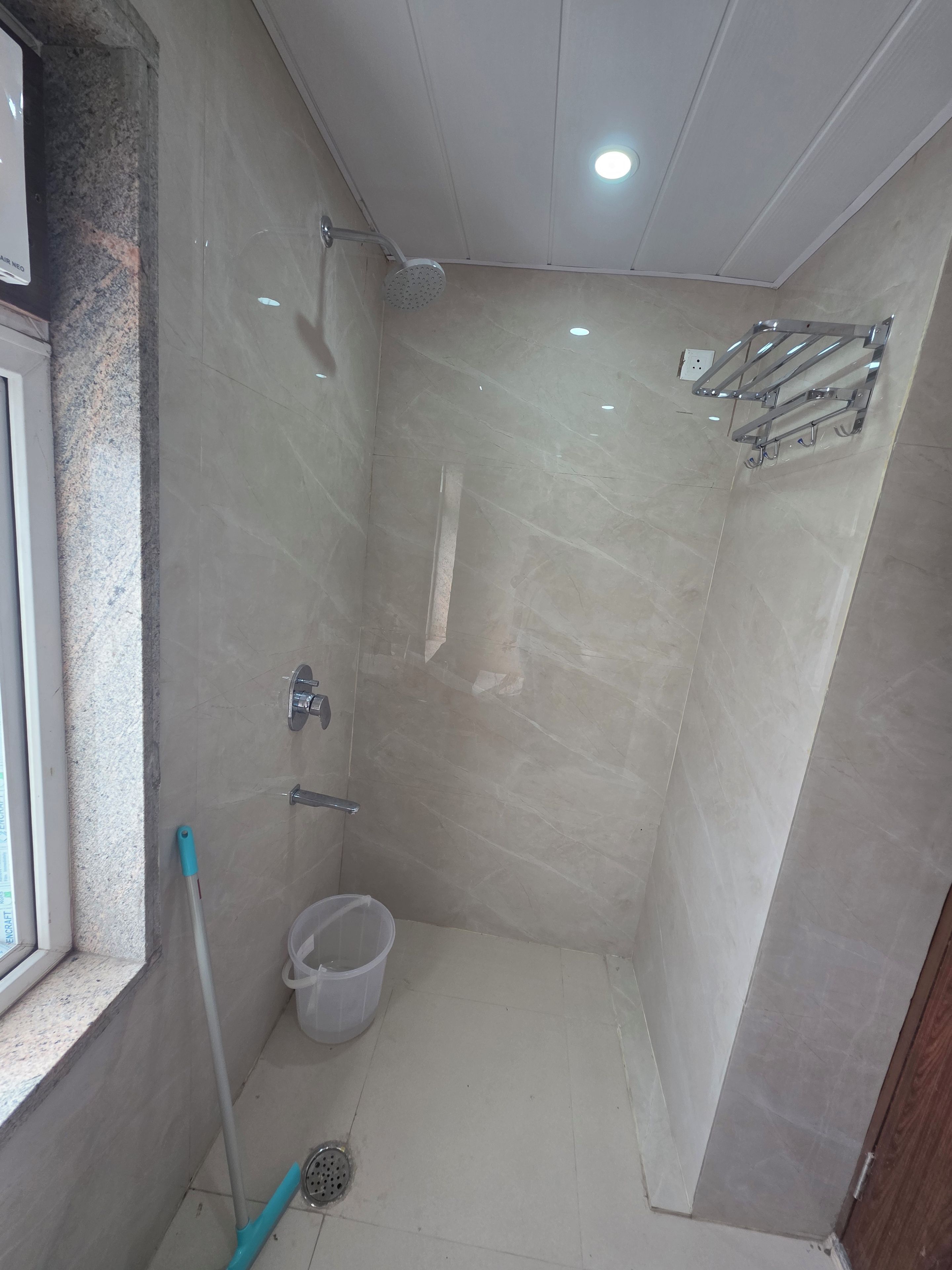 A small, tiled shower area with a handheld showerhead, a handheld mop, and a plastic bucket.