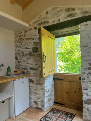 A rustic kitchen with a stone wall, a wooden cabinet, a small refrigerator, and a window with a view of greenery.
