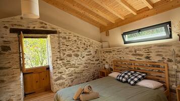 A bedroom with a sloped ceiling, wooden beams, a stone wall, a window, a bed with pillows, and a bedside table.