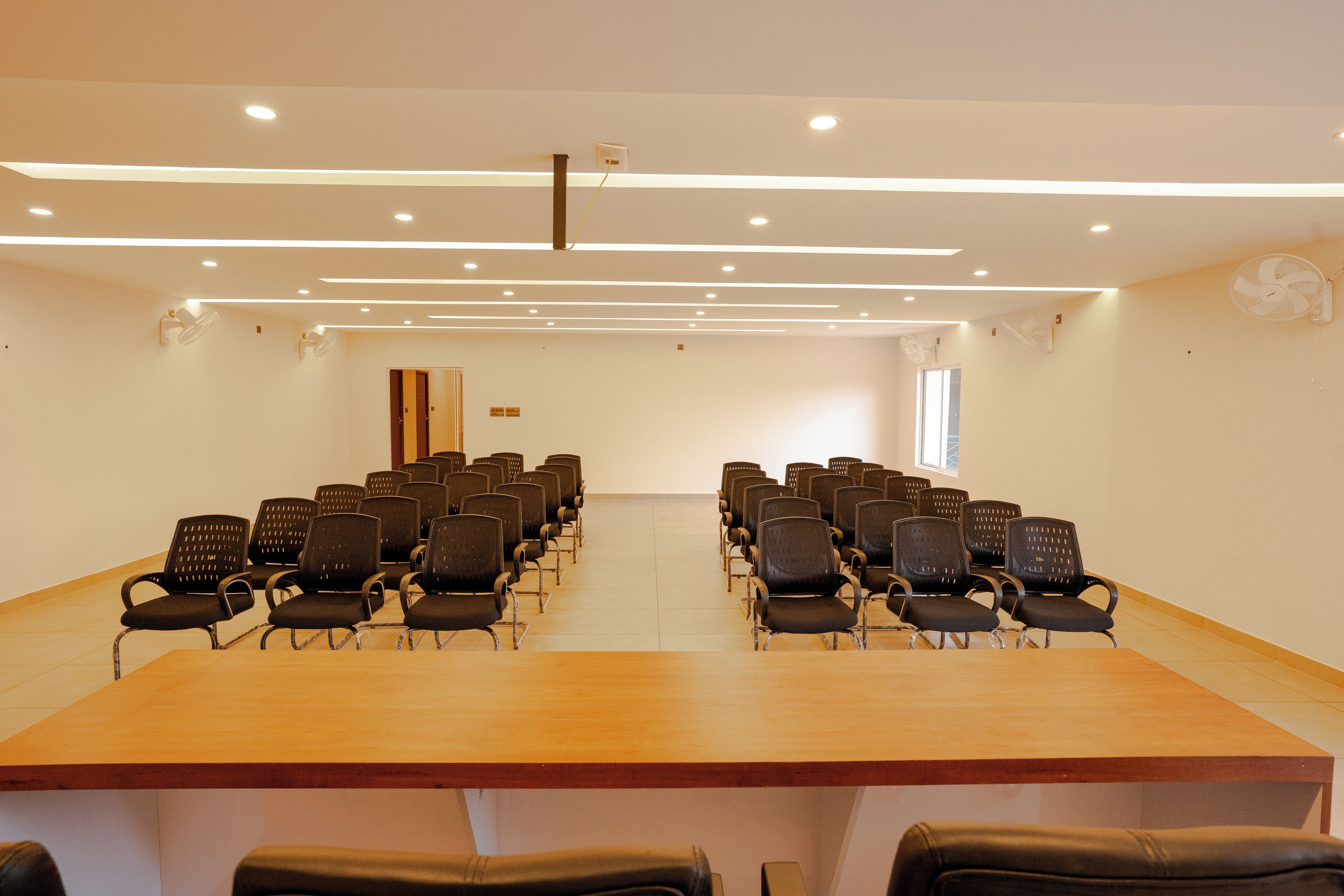 Un salón de conferencias con una larga mesa y filas de sillas, iluminación empotrada en el techo y un reloj en la pared.