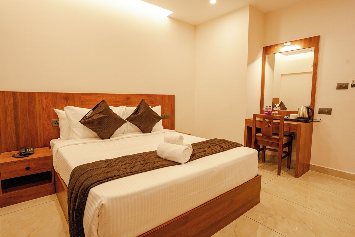 A hotel room with a wooden headboard, a bed with white linens, two pillows, a brown throw blanket, a wooden nightstand with a telephone, a desk with a chair, and a mirror on the wall.