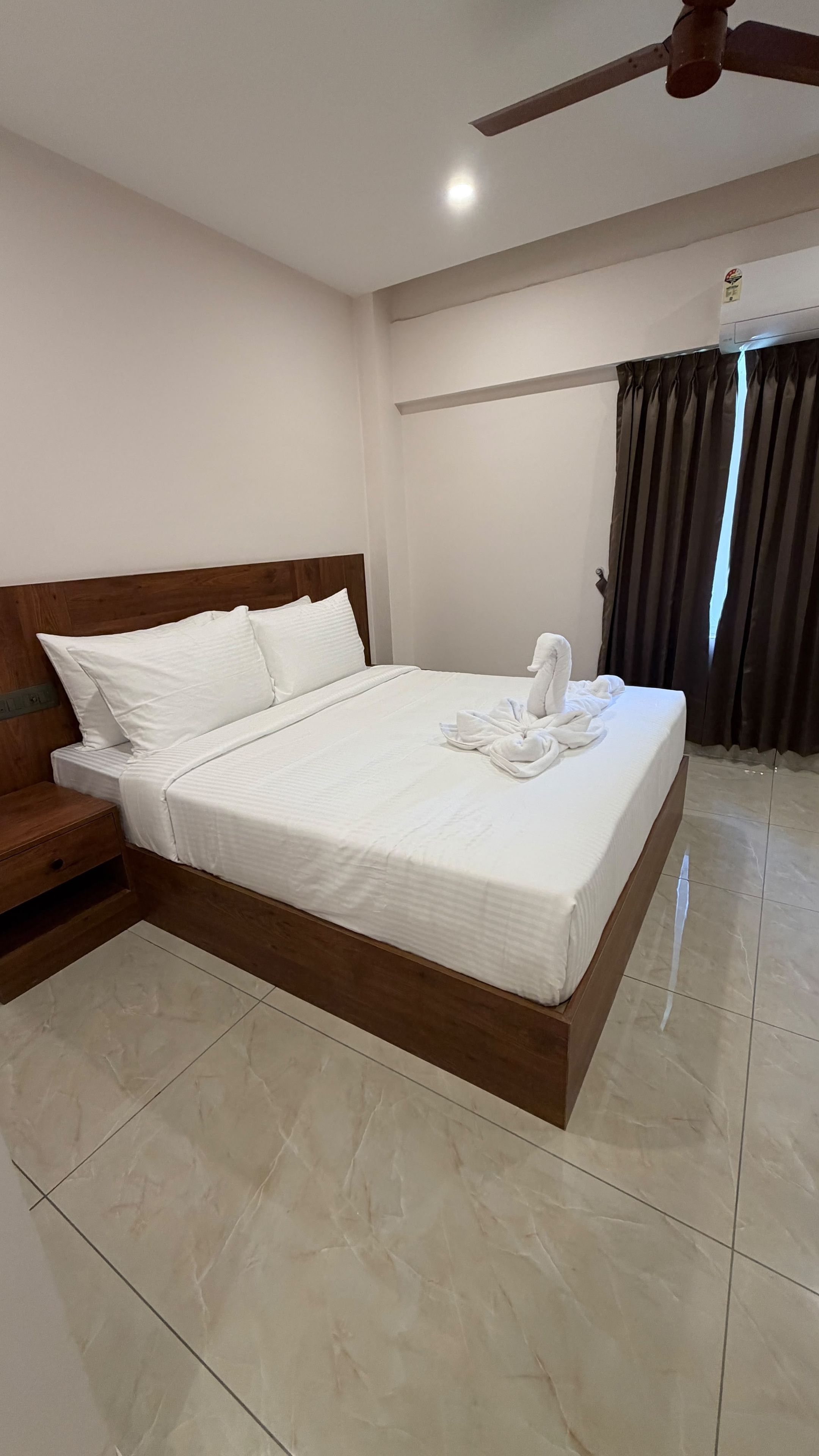 Una cama bien hecha con cabecera de madera, una mesita de noche pequeña y un ventilador de techo.