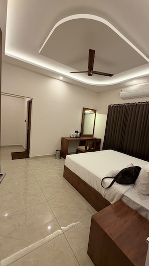 A hotel room with a bed, a desk with a mirror, a ceiling fan, and a window with curtains. - HAVENS BAY (Kumily)