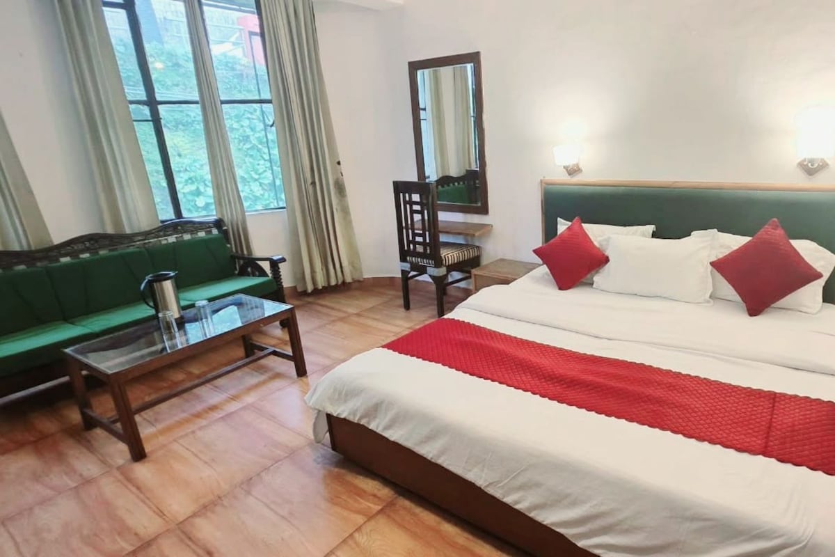 A hotel room with a bed, a green sofa, a wooden coffee table, a chair, a bedside table with a lamp, and a window with curtains.