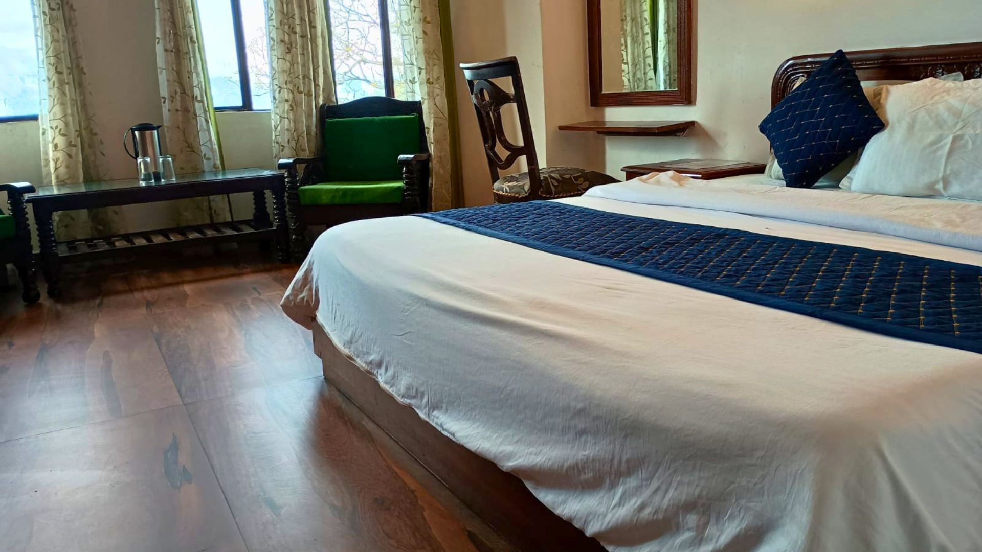 Habitación de hotel con una cama grande, un banco de madera, una silla y un ventanal con cortinas.
