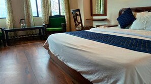 Quarto de hotel com uma cama grande, um banco de madeira, uma cadeira e uma janela com cortinas.