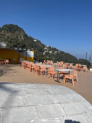 Uma área de estar externa com mesas e cadeiras, uma parede amarela e vista para uma colina com edifícios.