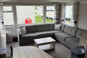 A modern living room with a grey sectional sofa, a wooden coffee table, and a view of a residential area through large windows.