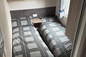 A compact bedroom with two beds, a small bedside table, and a window with curtains. - Cabin Sleeps 6 With Dishwasher (Skegness)