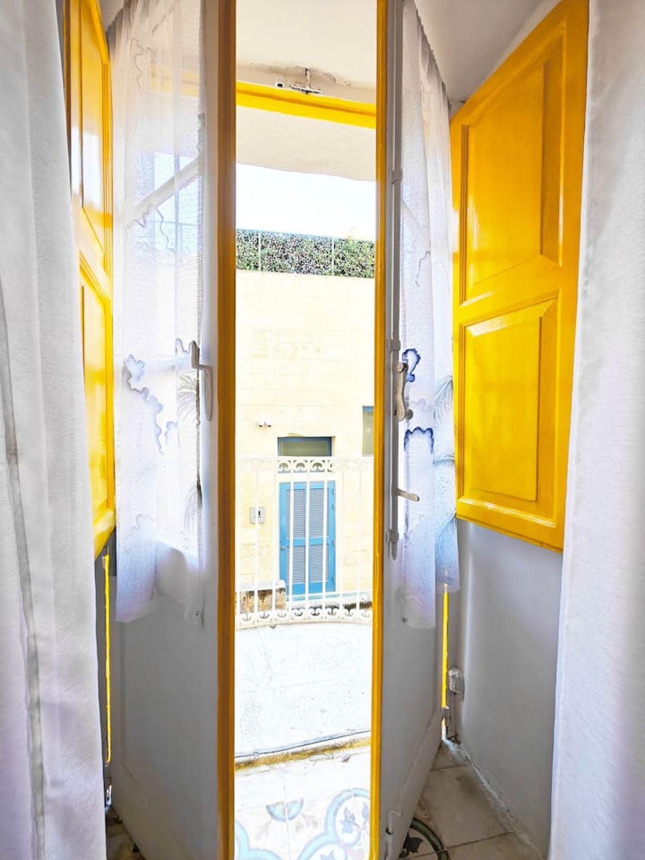 Een deuropening met een gele deur en een witte muur, die uitkomt op een buitenruimte met een blauwe deur en een balkon.