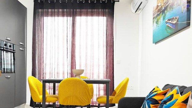 A modern living room with a grey sofa, colorful cushions, a dining table with yellow chairs, and a large window with curtains. - White tulip apartment (Safi)