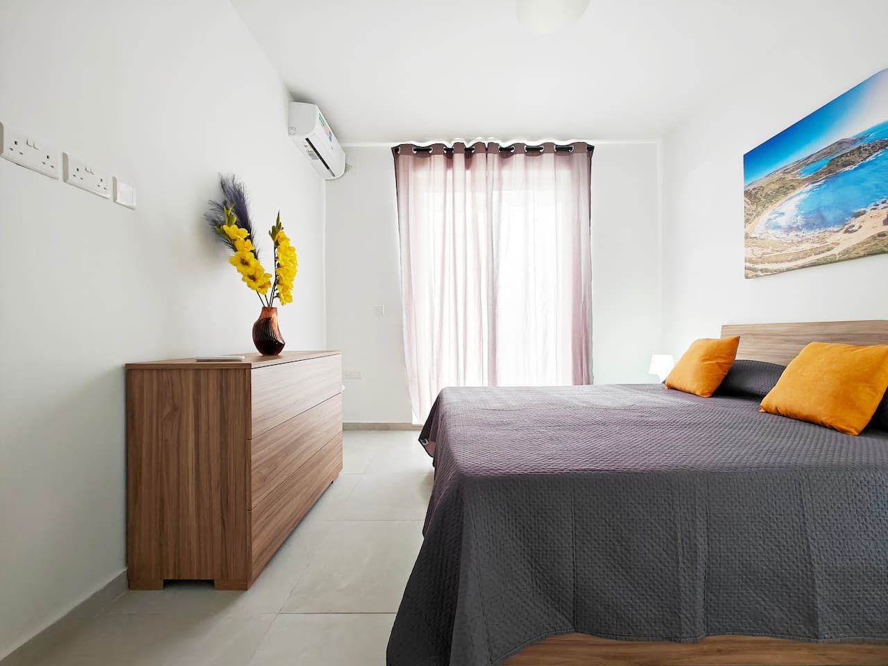 A bedroom with a bed, a wooden dresser, a vase with yellow flowers, a window with curtains, and a painting on the wall.