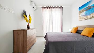 A bedroom with a bed, a wooden dresser, a vase with yellow flowers, a window with curtains, and a painting on the wall.