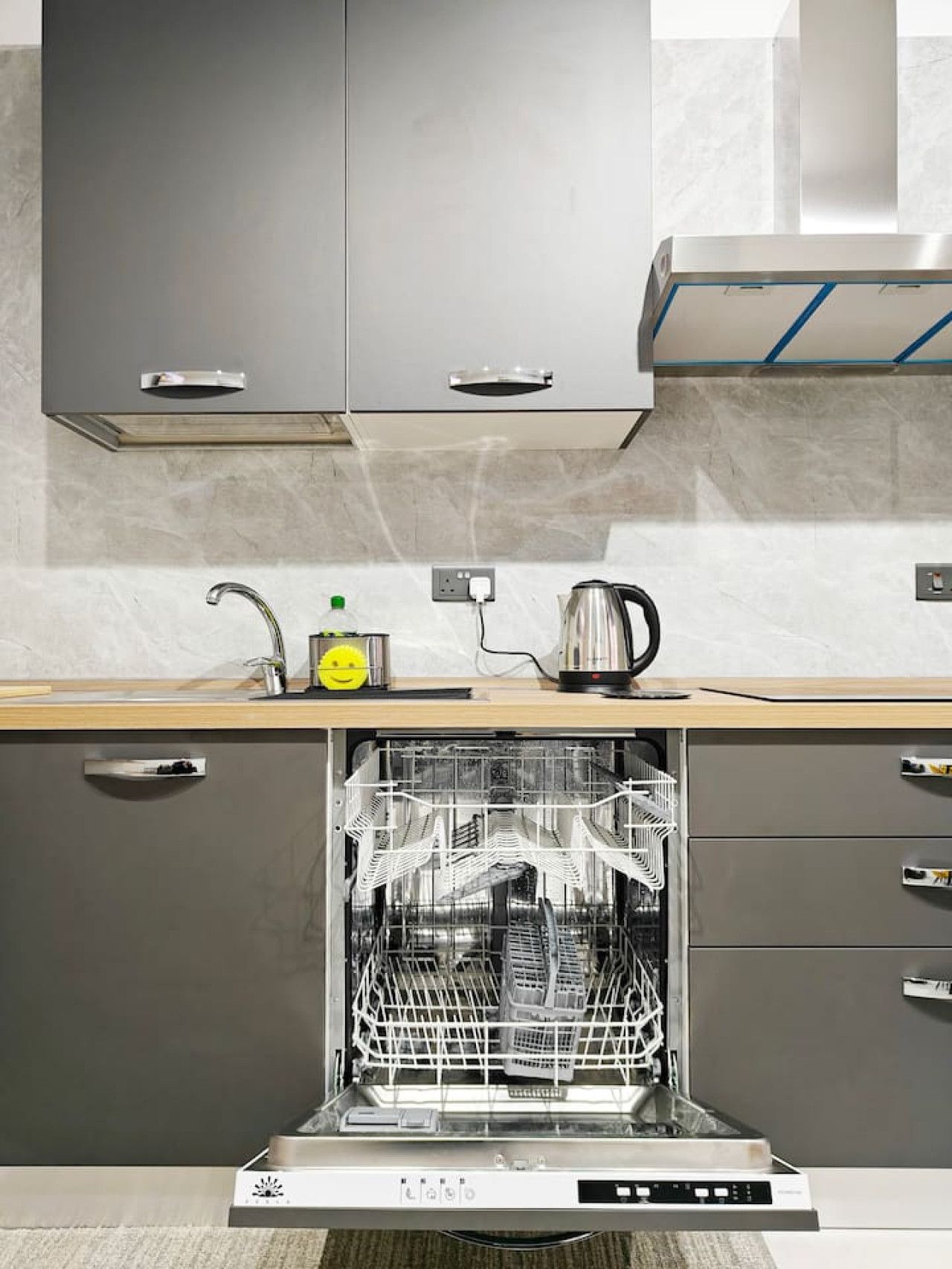 Una cucina moderna con mobili grigi, una lavastoviglie in acciaio inossidabile e un piano di lavoro con un bollitore e una bottiglia.