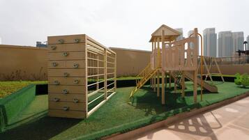 Un terrain de jeux équipé de structures d’escalade et d’un toboggan, installé sur une pelouse artificielle avec une vue sur le paysage urbain en arrière-plan.