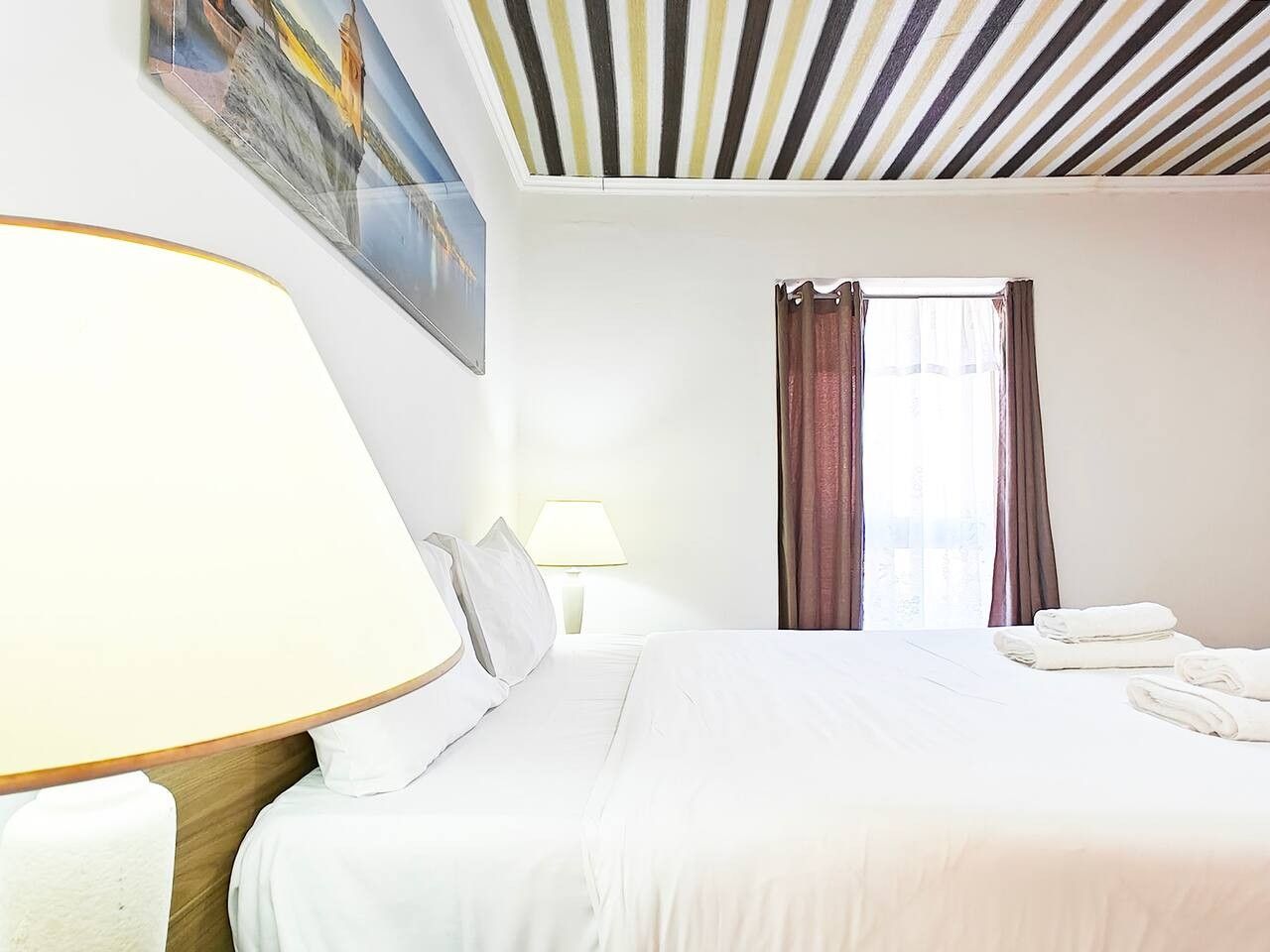 A hotel room with a bed, a lamp, a window with curtains, and a painting on the wall.