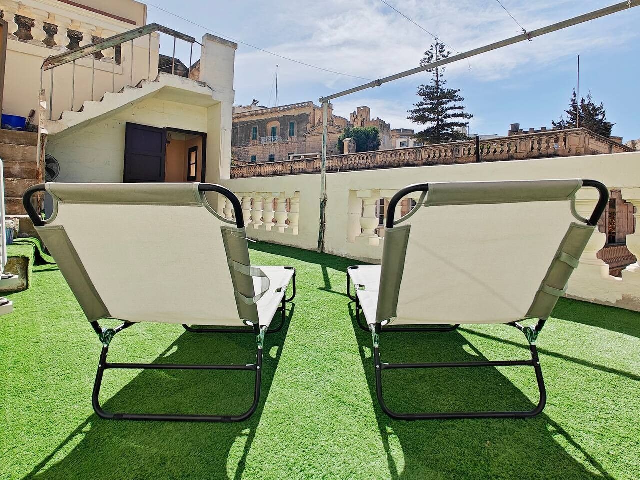 Two white lounge chairs with metal frames on a green artificial turf surface, with a building and a bridge in the background.