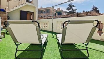 Two white lounge chairs with metal frames on a green artificial turf surface, with a building and a bridge in the background.