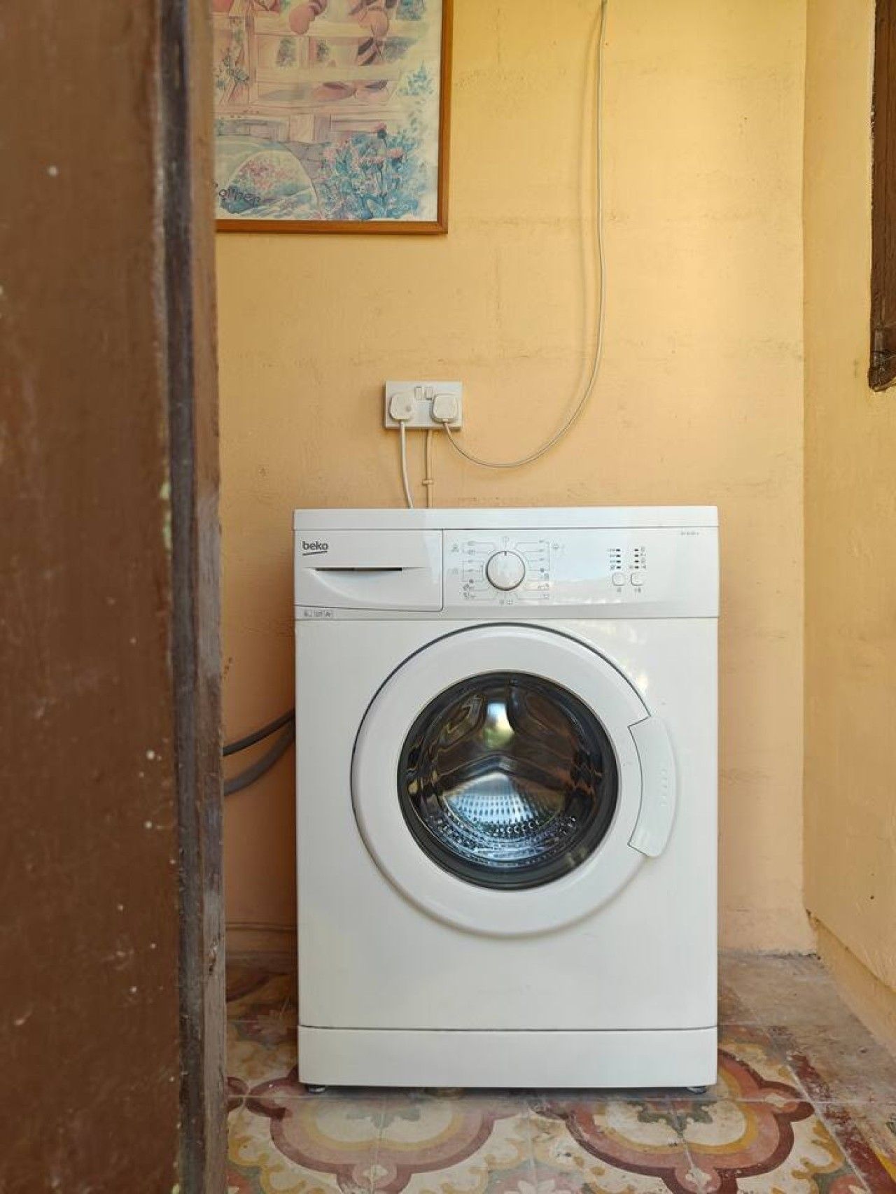 A front-loading washing machine in a room with a tiled floor and a wall-mounted electrical outlet.