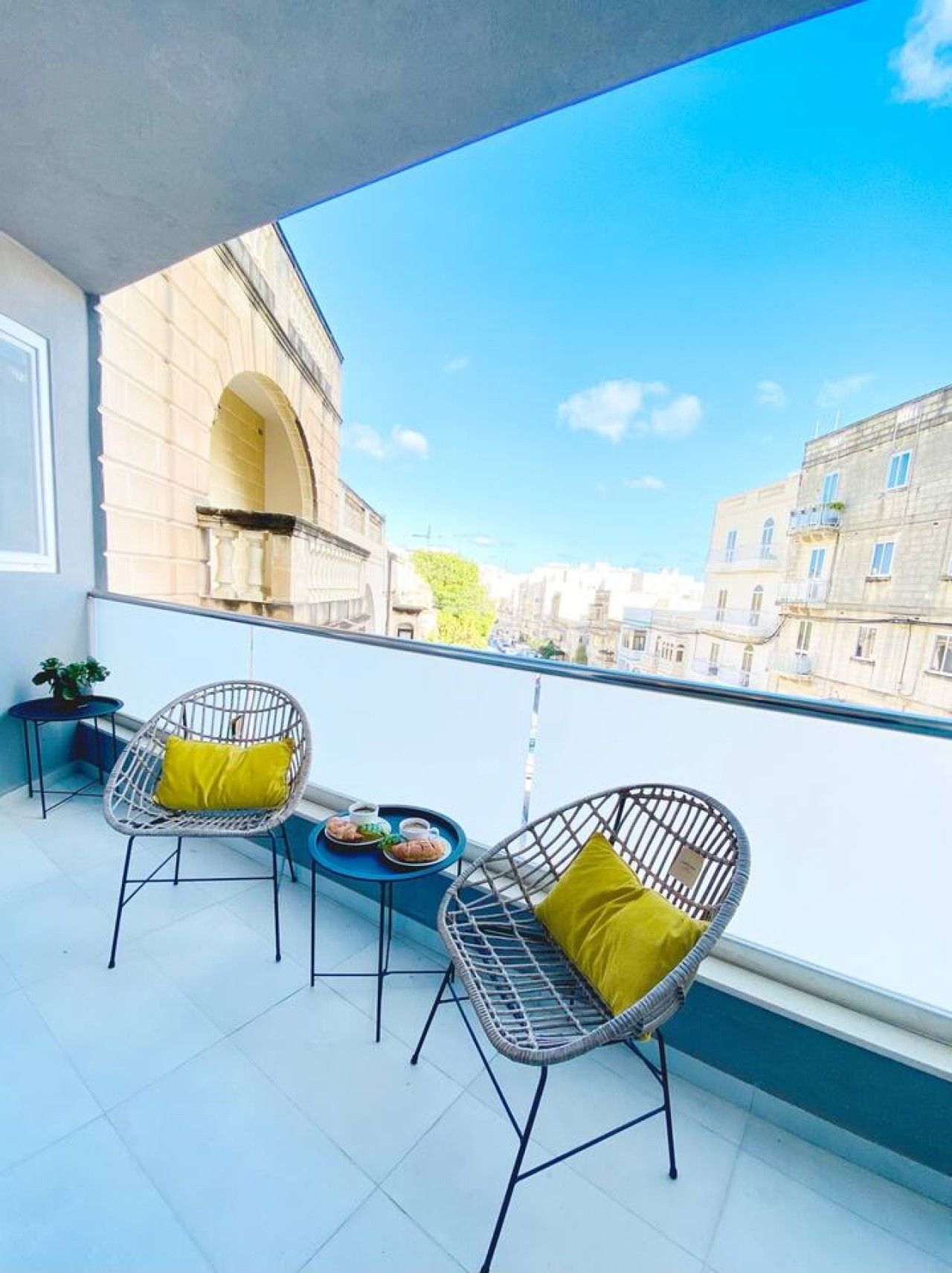 A balcony with two wicker chairs and a small table, overlooking a cityscape with buildings and a clear sky.