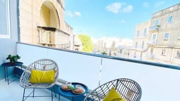 A balcony with two wicker chairs and a small table, overlooking a cityscape with buildings and a clear sky.