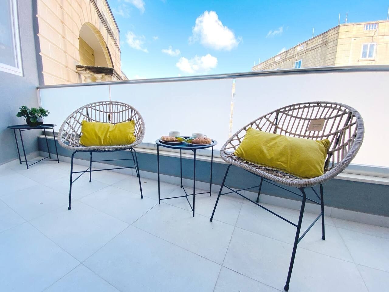 Une terrasse avec deux chaises en osier et une petite table, une plante en pot et un bâtiment en arrière-plan.