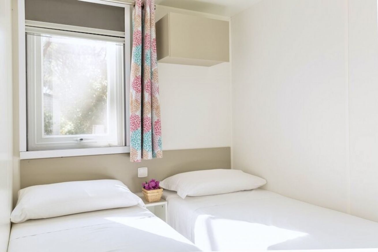 A bedroom with two beds, a window with a curtain, and a small bedside table with a flower vase.