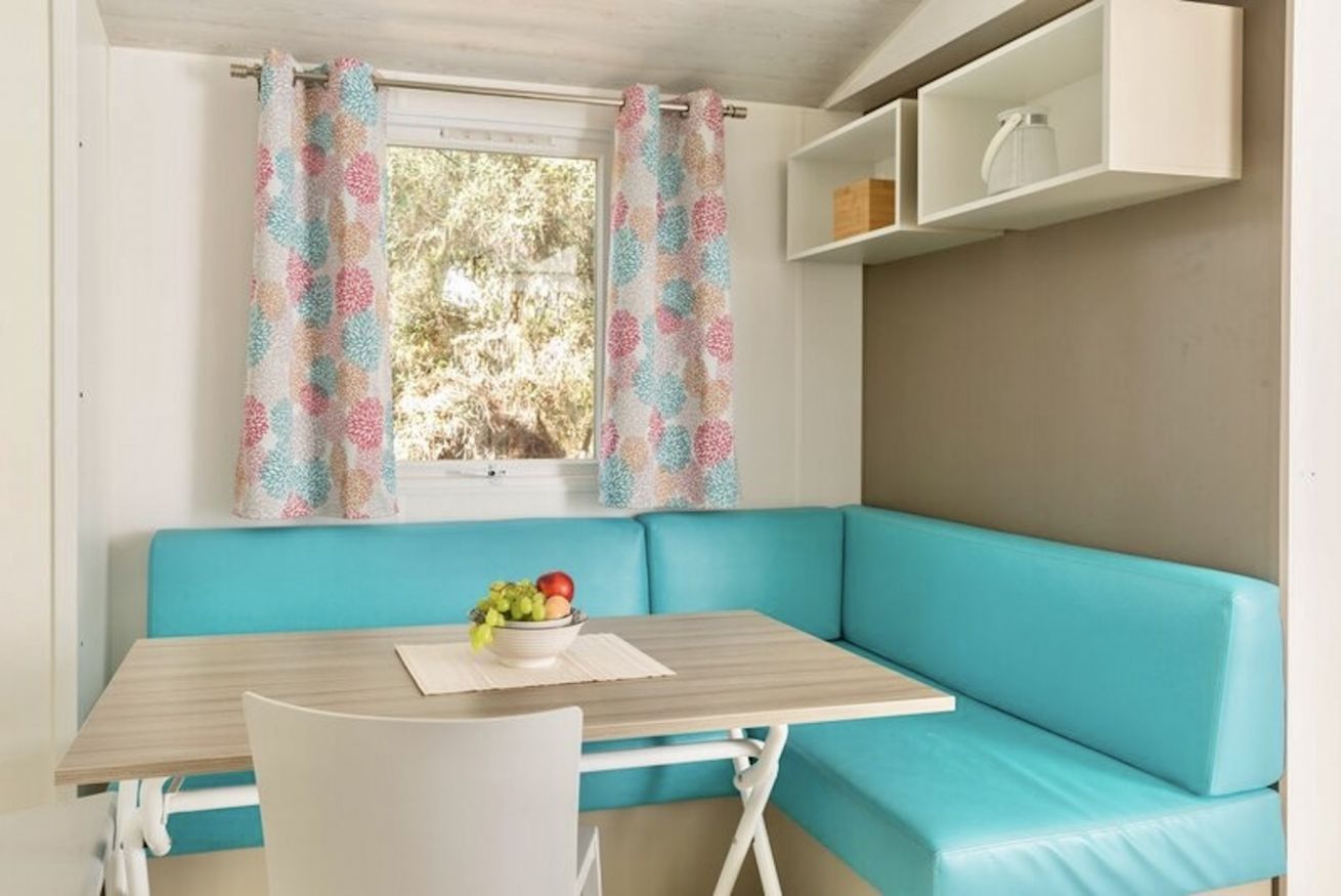 A room with a teal sofa, a small table with a bowl of fruit, and a window with patterned curtains.