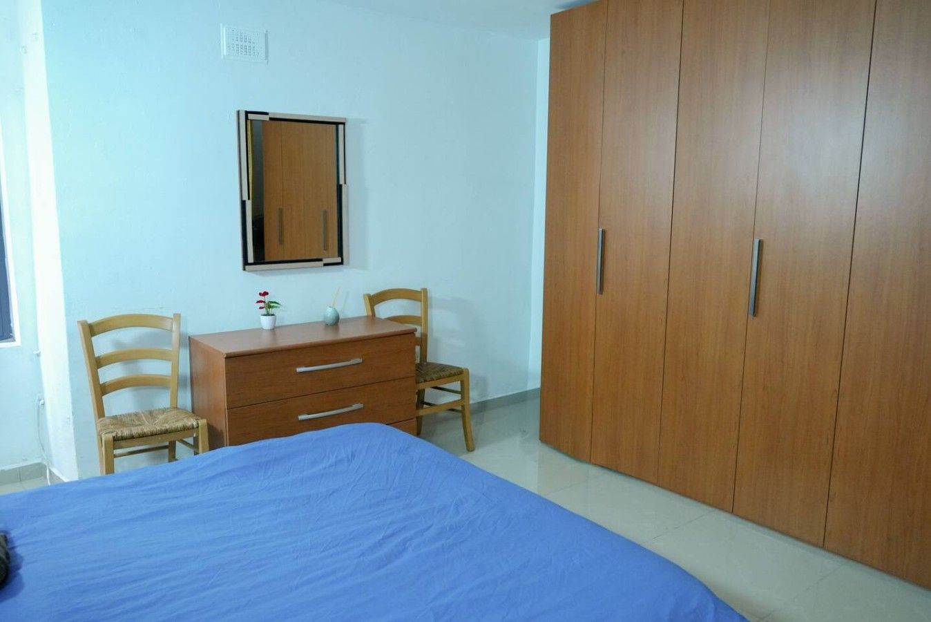 Una camera da letto con un letto, un'armadiatura in legno, una cassettiera, due sedie e uno specchio a parete.