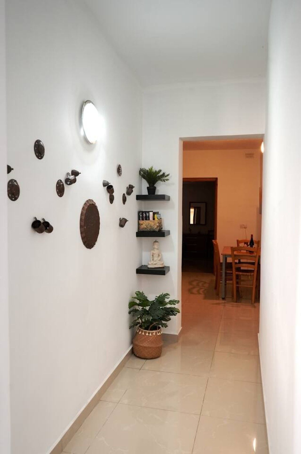Un corridoio con parete bianca decorata con oggetti ornamentali, una pianta in vaso e un'area pranzo con mobili in legno.