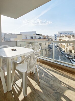 A balcony with a table and chairs, a view of a street with parked cars, and buildings in the background. - Black Diamond apartment (Żabbar)
