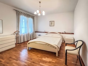 A bedroom with a bed, bedside tables, a chair, a dresser, a window with curtains, and a chandelier. - La Casa del Nespolo (Torrita di Siena)